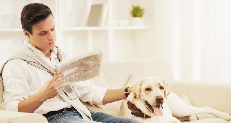 Man reading a newspaper on the sofa while he pets his dog who sits next to him